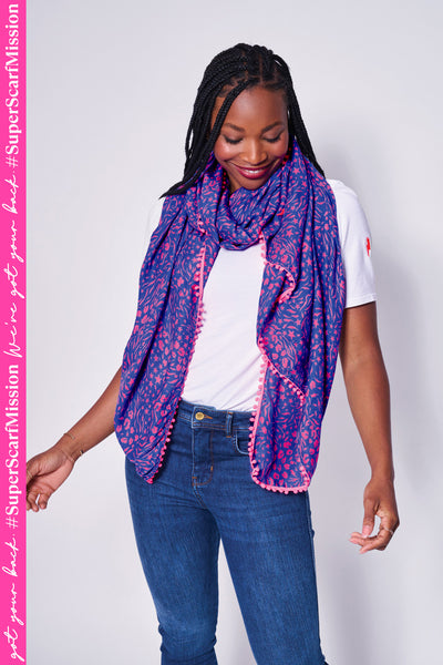 A lady wearing a blue with pink mixed animal and lightning bolt print scarf with a pink pom pom trim
