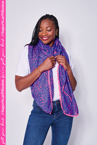 A lady cosying up in a blue with pink mixed animal and lightning bolt print scarf with a pink pom pom trim