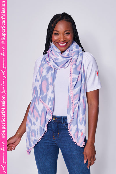 A lady wearing a white with pale blue and pale pink leopard ikat and lightning bolt print scarf with a pink pom pom trim