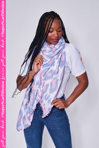 A lady wearing a white with pale blue and pale pink leopard ikat and lightning bolt print scarf with a pink pom pom trim