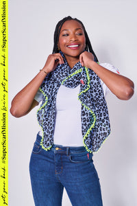 A lady wearing a soft blue with black floral leopard and lightning bolt print scarf with a neon yellow pom pom trim