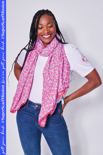 A lady wearing a soft blue with coral floral leopard and lightning bolt print scarf with a coral pom pom trim 
