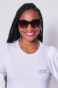 A lady smiling wearing black cat eye sunglasses with a white Scamp & Dude T-shirt and gold jewellery