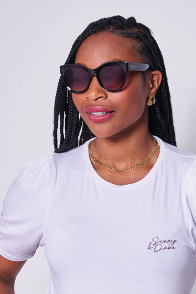 A lady wearing black cat eye sunglasses with a white Scamp & Dude T-shirt and gold jewellery