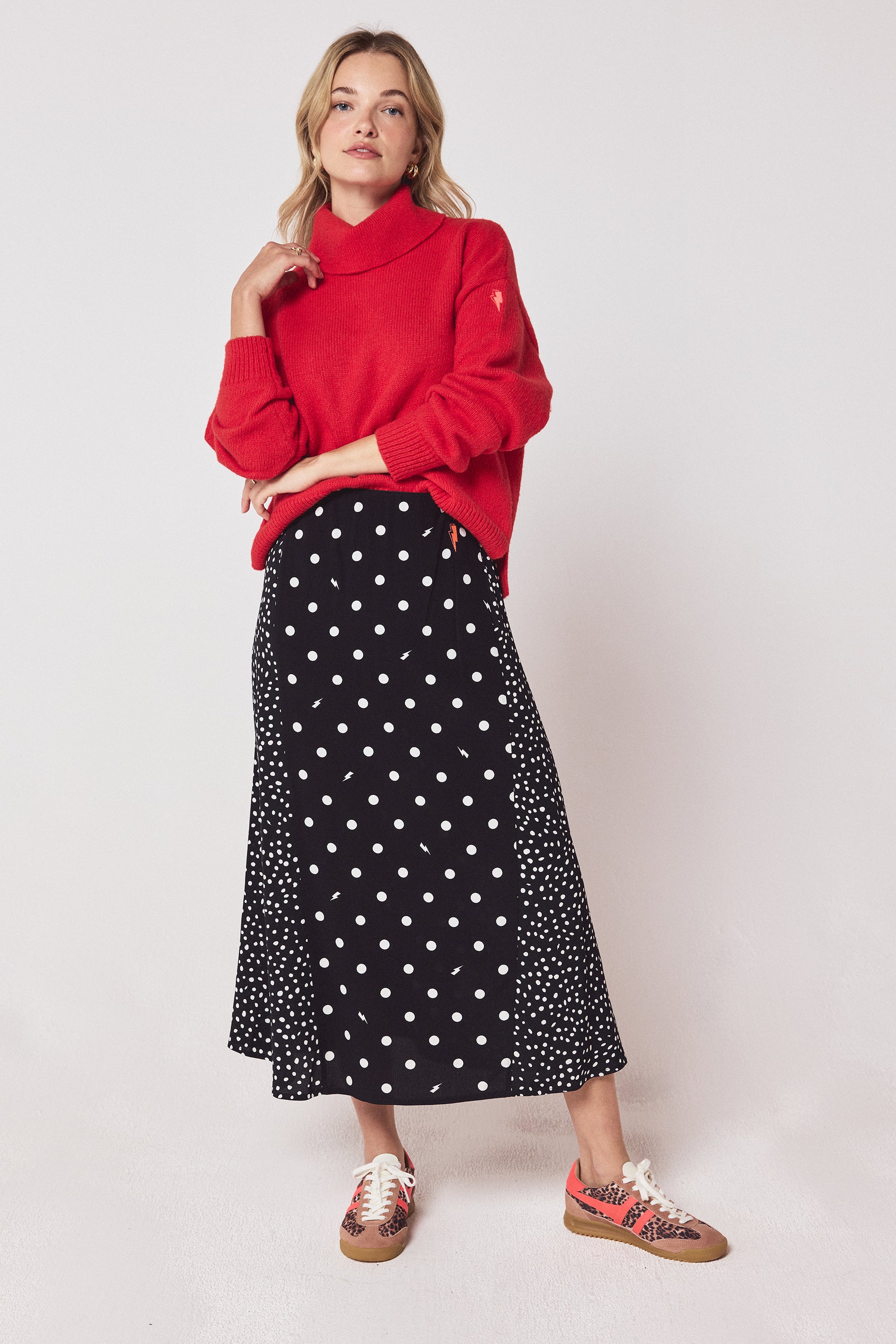 Scamp & Dude Black with White Polka Dot Bolt Midi Skirt | Model wearing a red roll neck jumper with a black polka dot midi skirt and cheetah print trainers.