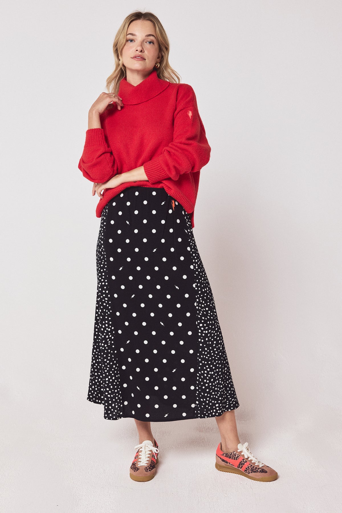 Scamp & Dude Black with White Polka Dot Bolt Midi Skirt | Model wearing a red roll neck jumper with a black polka dot midi skirt and cheetah print trainers.