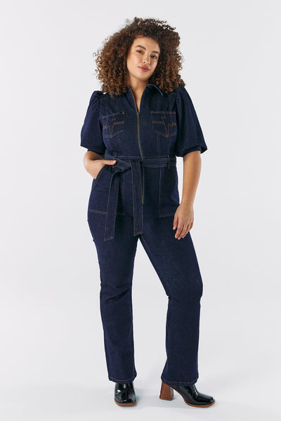 Scamp and Dude Rinse Wash Denim Flared Leg Jumpsuit | Model with curly hair wearing dark wash denim jumpsuit with high heeled boots