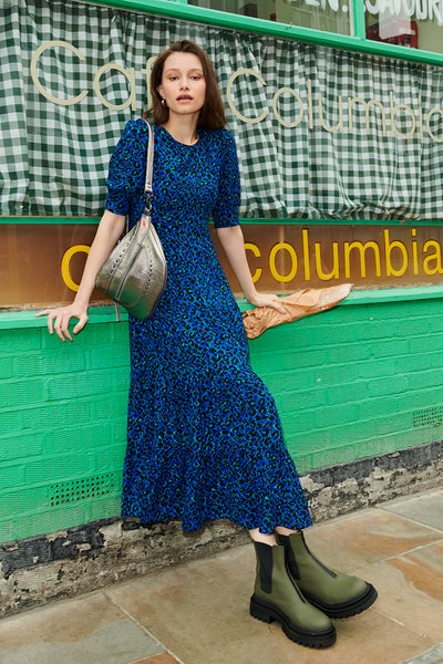 Scamp and Dude Blue with Green and Black Small Shadow Leopard Maxi Dress | Model wearing long blue dress leaning against a green shop front with a baguette in hand and green boots