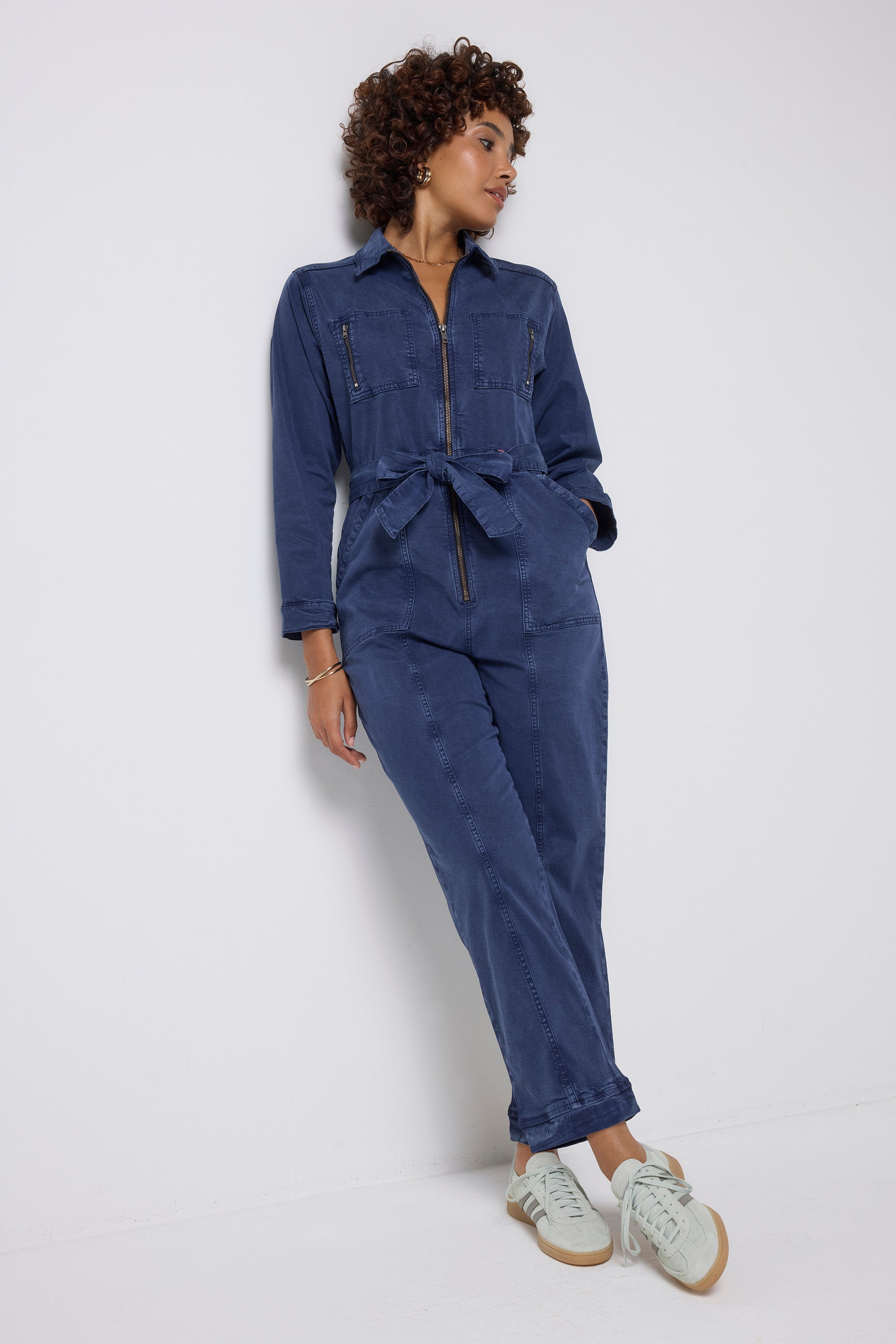 Scamp and Dude Ink Blue Tie Front Boiler Jumpsuit | Model wearing a ink blue jumpsuit featuring a zip front and tie waist, paired with white trainers.