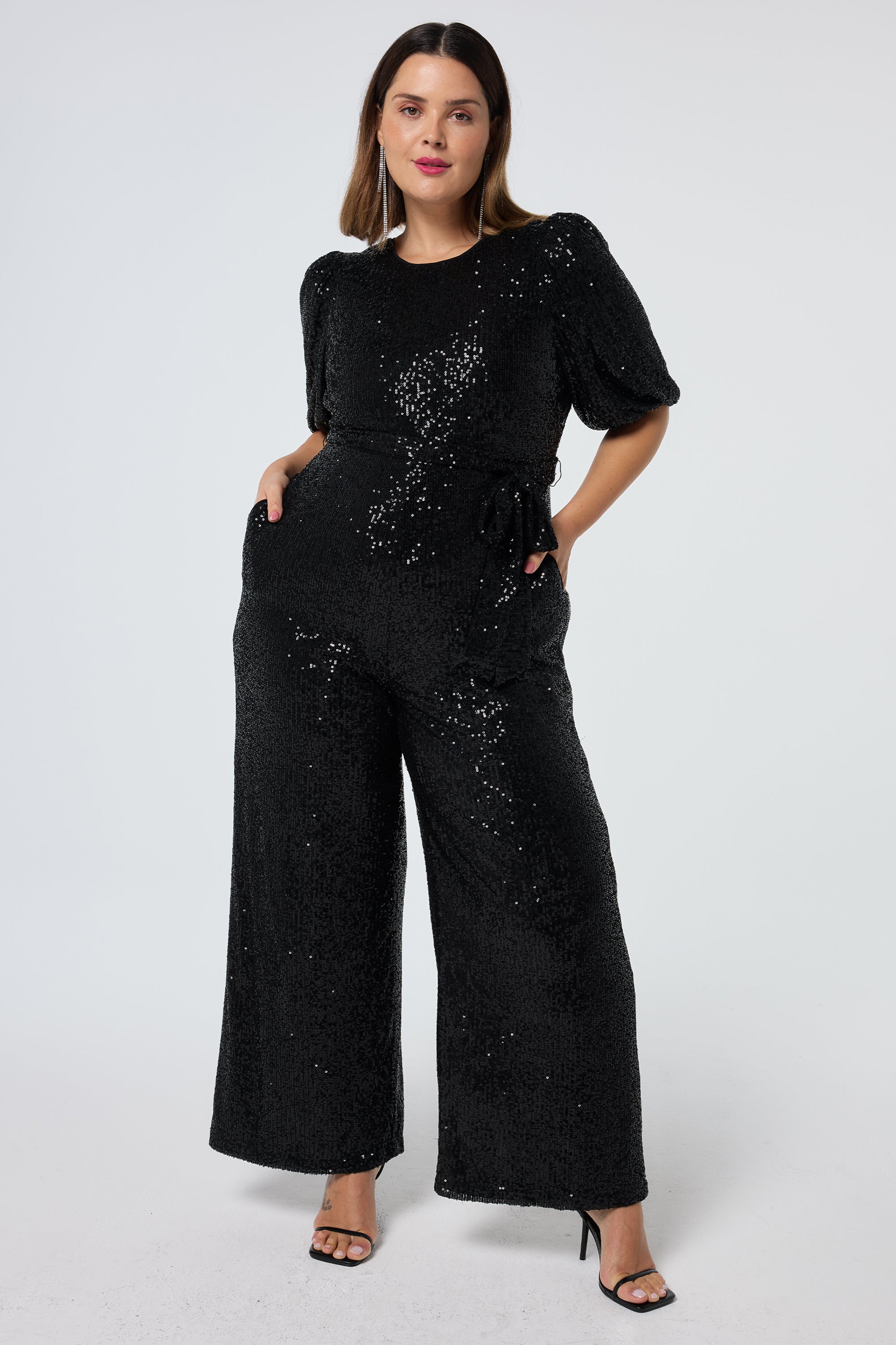 Scamp and Dude Black Sequin Puff Sleeve Wide Leg Jumpsuit | Model wearing a black wide leg sequin jumpsuit with a tie waist feature and black heels.