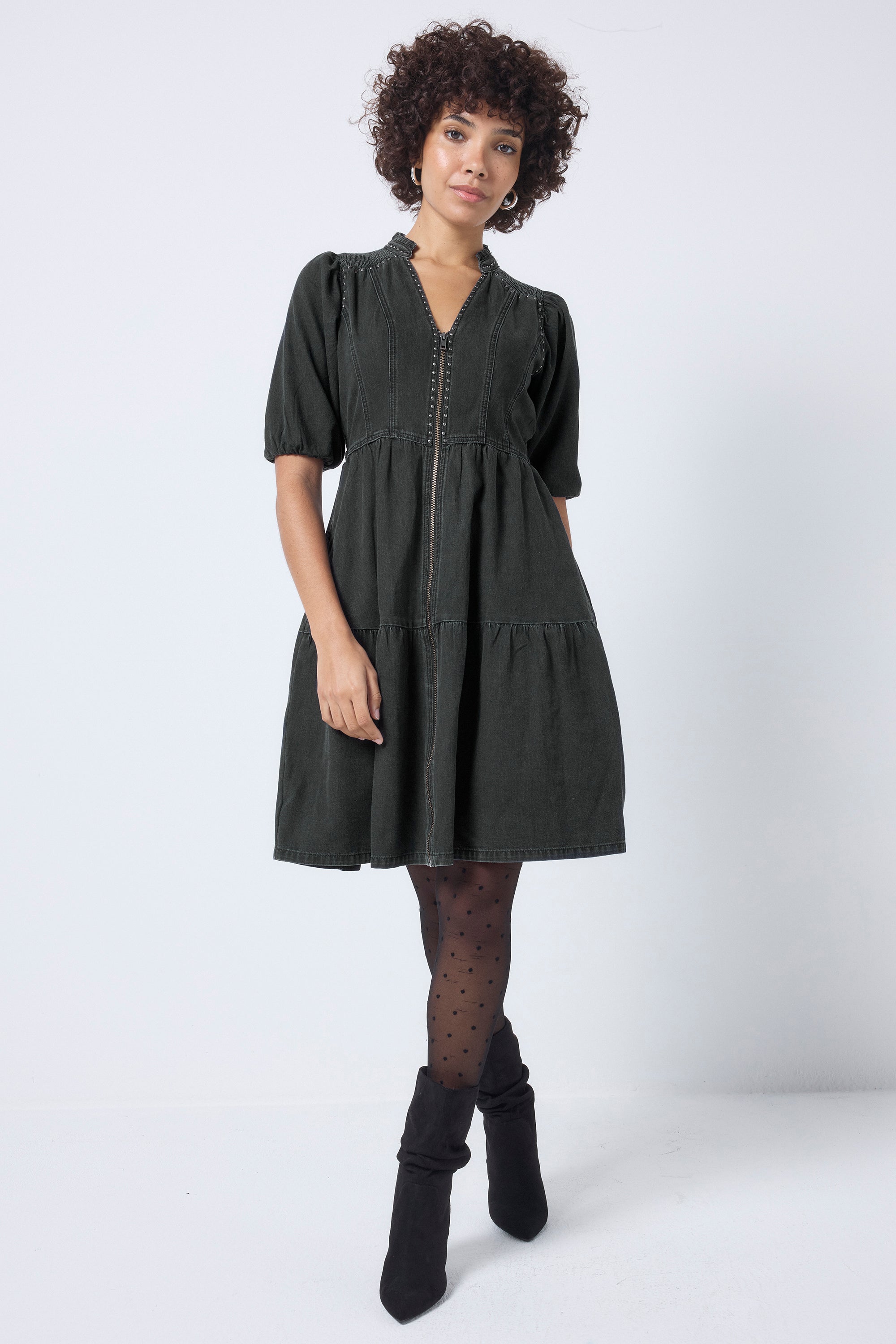 Scamp and Dude Washed Black Studded Zip Detail Tiered Short Denim Dress | Model wearing short denim zip dress with stud detail. Paired with black polka dot tights and black boots.