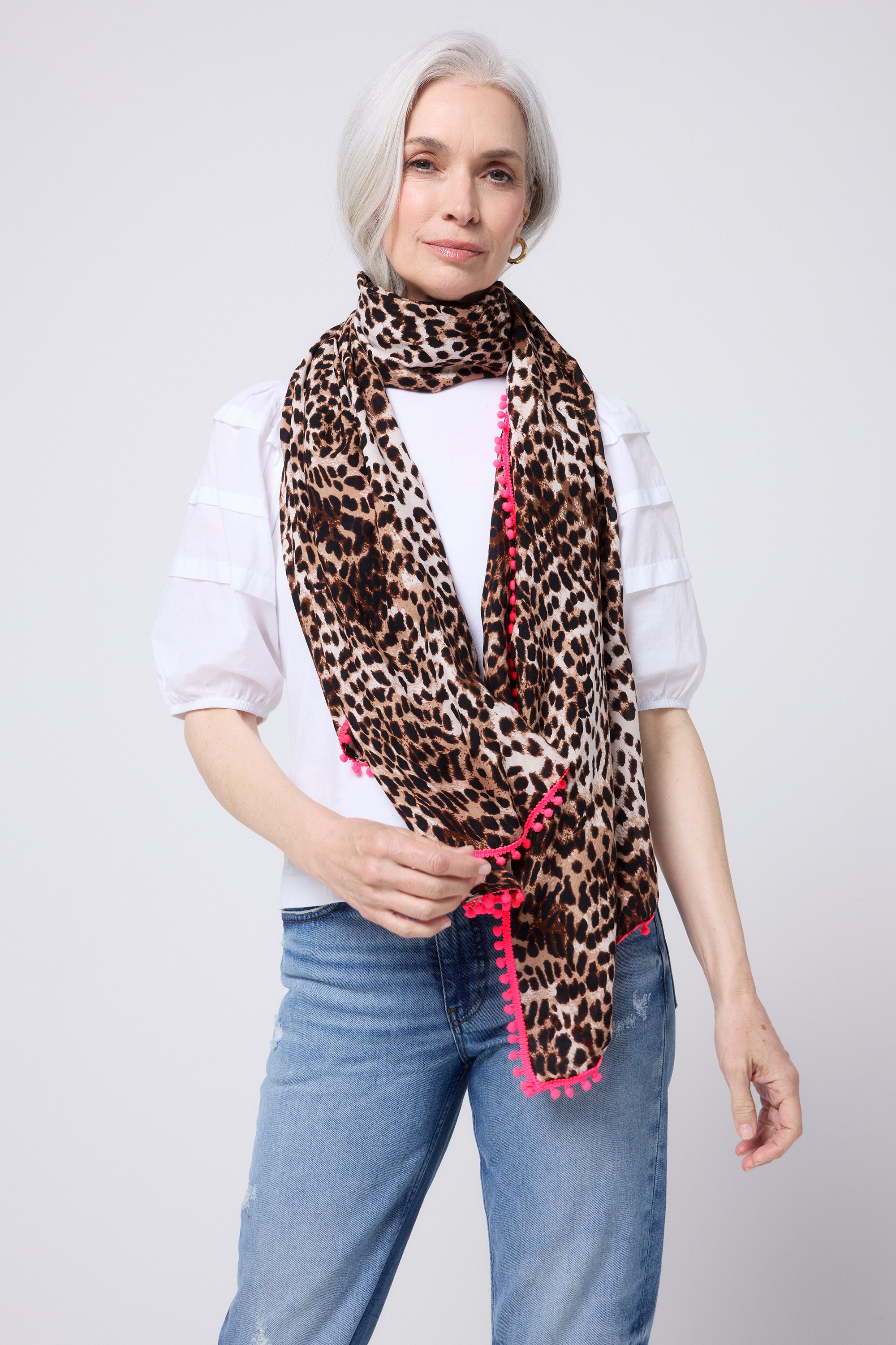 Scamp and Dude Neutral with Black Cheetah Charity Super Scarf | Model wearing a pair of blue jeans with a white t shirt and neutral scarf with all over cheetah print and neon coral pom pom detail.
