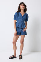 Scamp and Dude Indigo Button Through Denim Top | Model wearing a blue denim button through top with puff sleeves. Worn with blue denim shorts and black sandals.