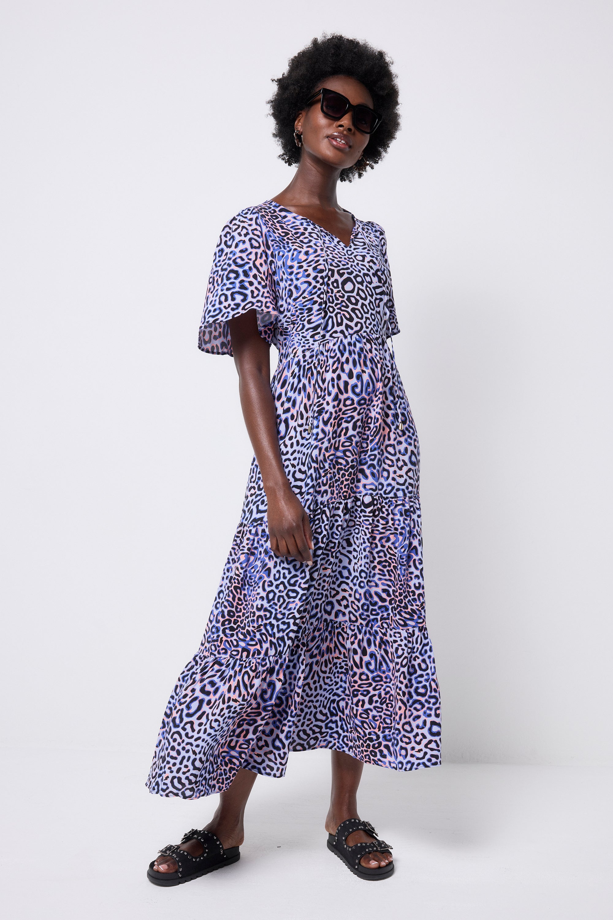 Scamp and Dude Lilac with Black and Pink Leopard Ray Tie Front Maxi Dress | Model wearing a lilac leopard ray print maxi dress with a v-neck and tie front detail. Paired with black studded sandals and tortoiseshell sunglasses.