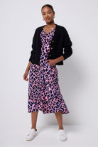 Scamp and Dude Black Cardigan | Model wearing a pink with blue and black leopard print dress with a black cardigan worn over it. Paired with white trainers and gold jewellery.