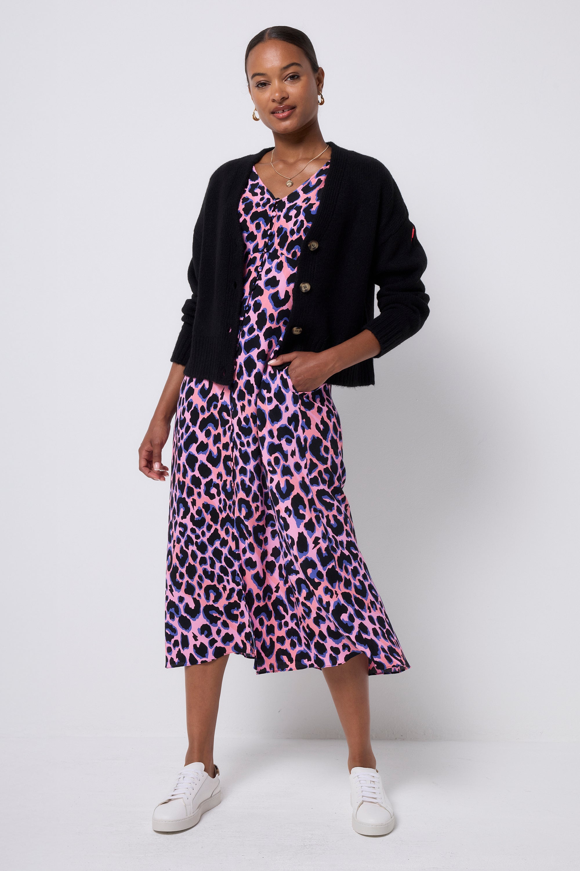Scamp and Dude Black Cardigan | Model wearing a pink with blue and black leopard print dress with a black cardigan worn over it. Paired with white trainers and gold jewellery.