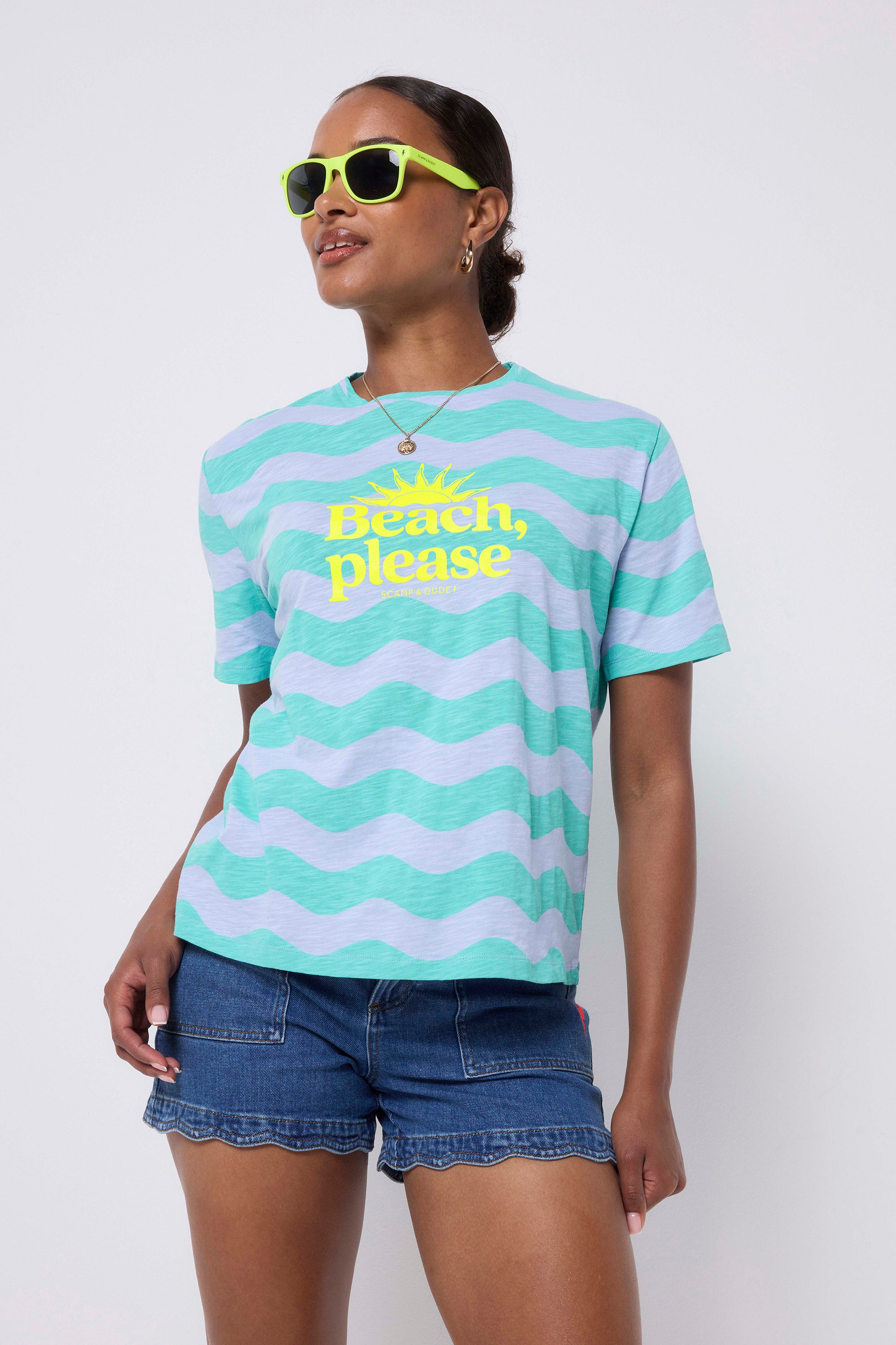 Scamp and Dude Blue with Green Wave Stripe Beach Please T-Shirt | Model wearing a pair of blue scallop denim shorts with a green and blue wave stripe t-shirt that features a yellow "Beach, please" and sun graphic print. Worn with neon yellow sunglasses and white trainers.