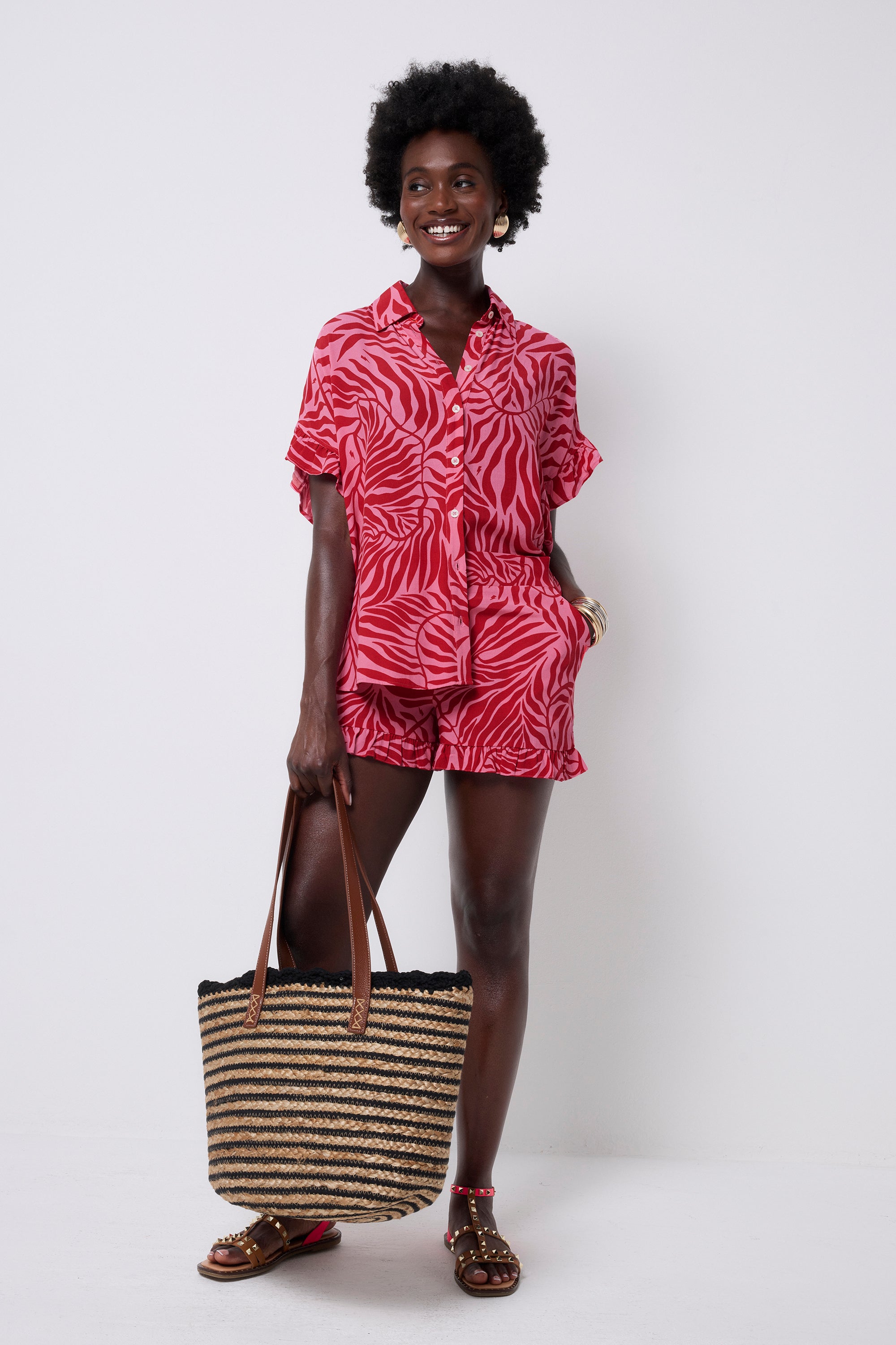 Scamp and Dude Red with Pink Palm Frill Sleeve Shirt | Model wearing a pink with red palm print button front shirt. Worn with a matching pair of shorts and sandals. She is holding a straw bag with black stripes.