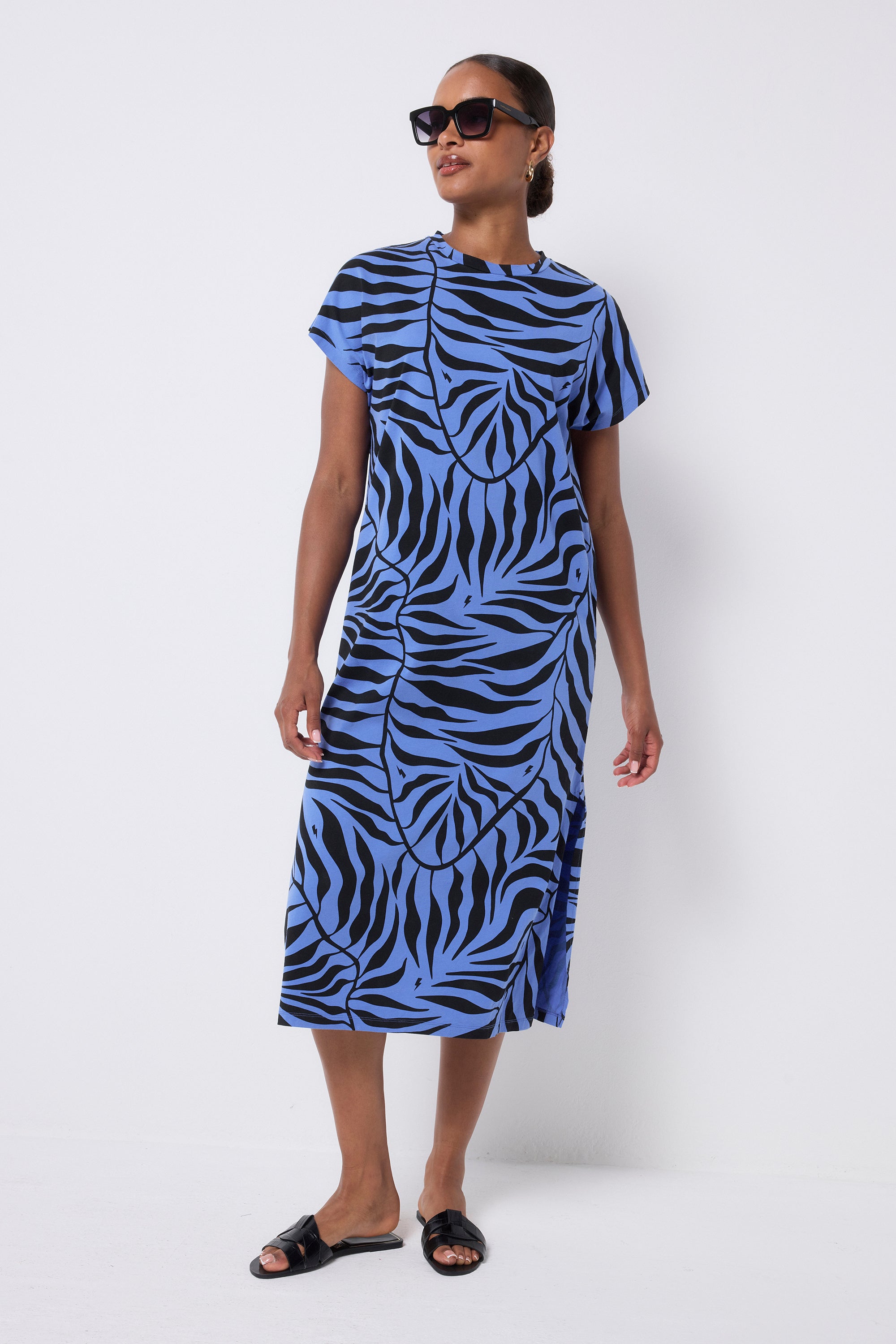 Scamp and Dude Blue with Black Palm Midi T-Shirt Dress | Model with her hair in a low bun wearing a blue crew neck t-shirt dress with black palm print. Paired with black sandals and sunglasses.
