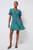 Scamp and Dude Green with Black Leopard Ray Short Skater Dress | Model wearing a green short dress with a v neck and puff sleeves. The dress has a black leopard ray print, it is worn with black sandals.