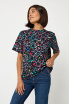 Scamp and Dude Grey with Rainbow Shadow Leopard Print T-Shirt | Model with short dark hair wearing a short sleeve grey t shirt that has rainbow leopard print all over. Worn with blue jeans.
