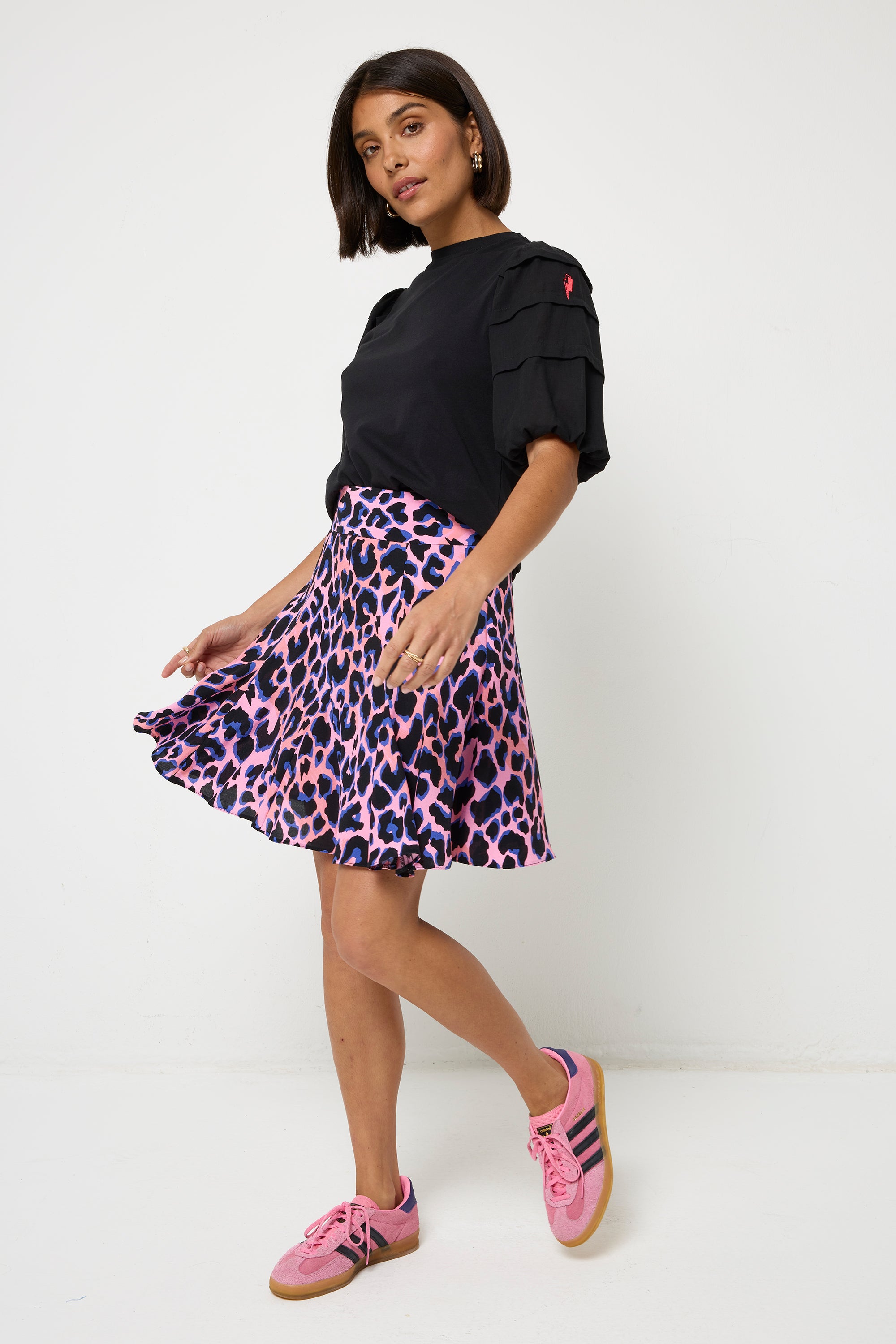 Scamp and Dude Pink with Blue and Black Shadow Leopard Skater Skirt | Model with short dark hair wearing a black puff sleeve t shirt with a pink and blue leopard print short skirt. Worn with pink trainers.