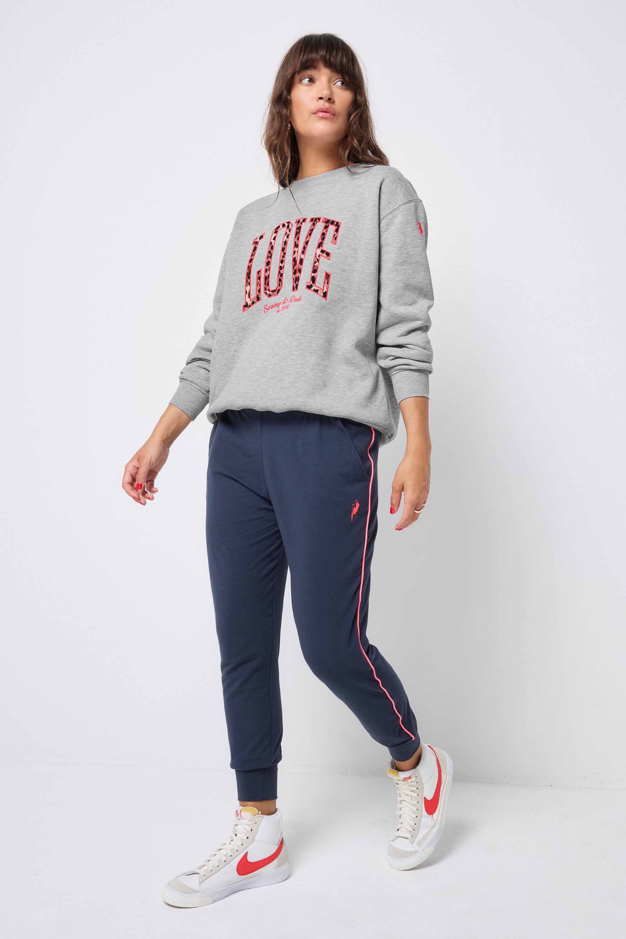 Scamp and Dude Navy Slouch Joggers | Model wearing a pair of navy joggers with neon coral piping detail, worn with a grey sweatshirt and white high top trainers.
