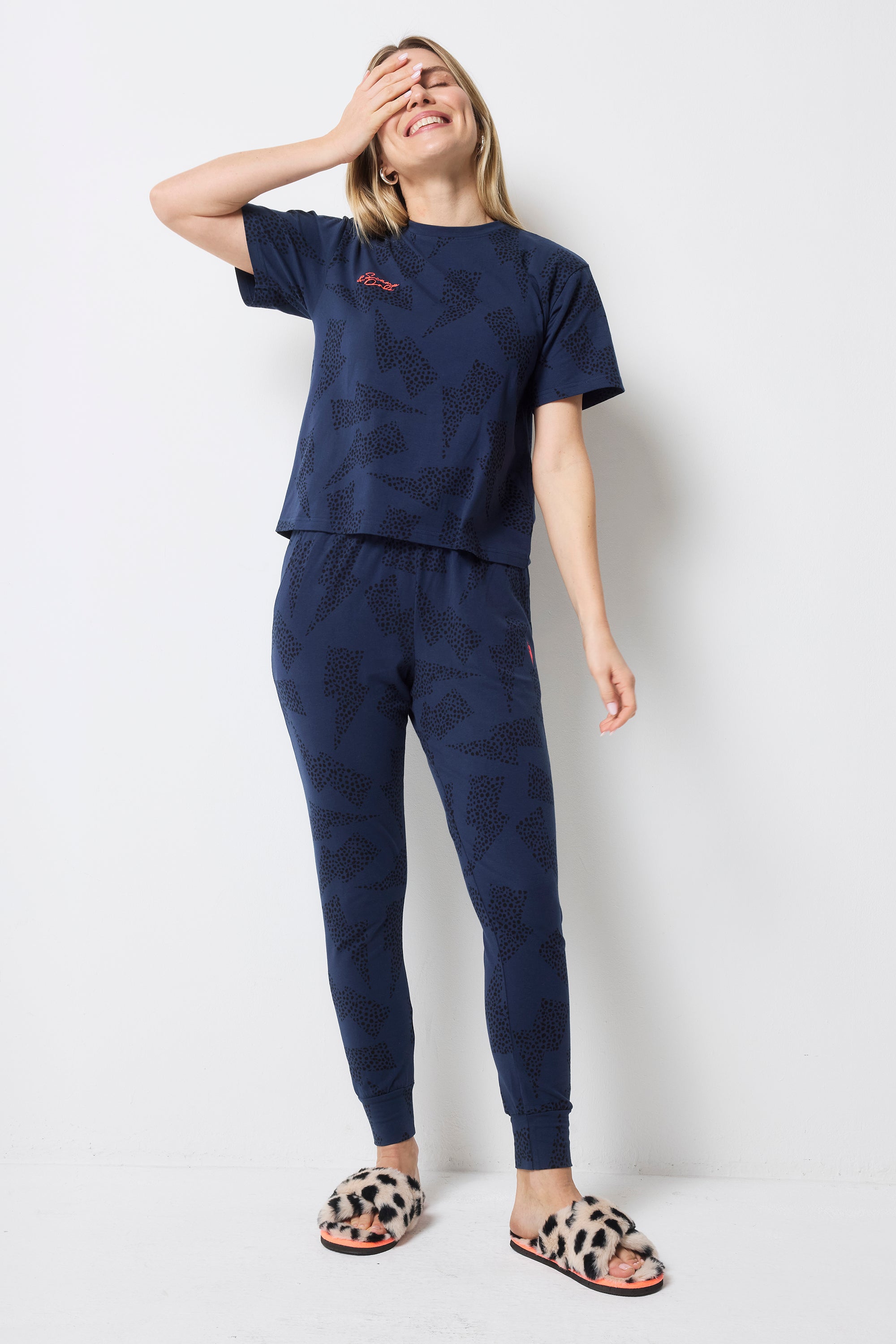 Scamp and Dude Navy with Black Cheetah and Lightning Bolt Print Pyjamas | Model wearing a pair of navy cheetah and lightning bolt print pyjamas with cream cheetah print slippers.