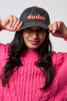 Scamp & Dude Washed Black Duder Cap | Model wearing a black cap with 'duder' text with a pink knitted jumper, posing against a plain white background.
