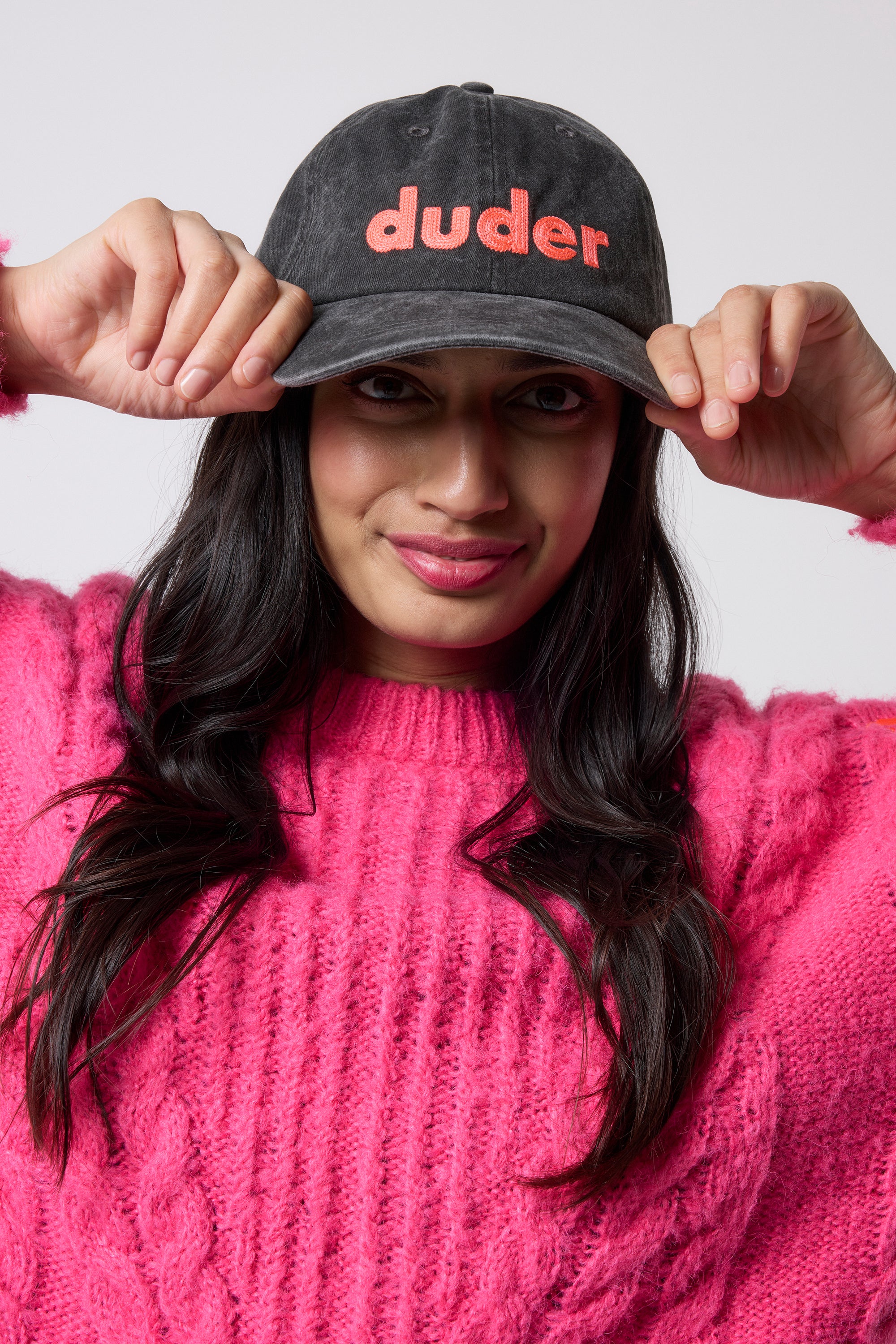 Scamp & Dude Washed Black Duder Cap | Model wearing a black cap with 'duder' text with a pink knitted jumper, posing against a plain white background.