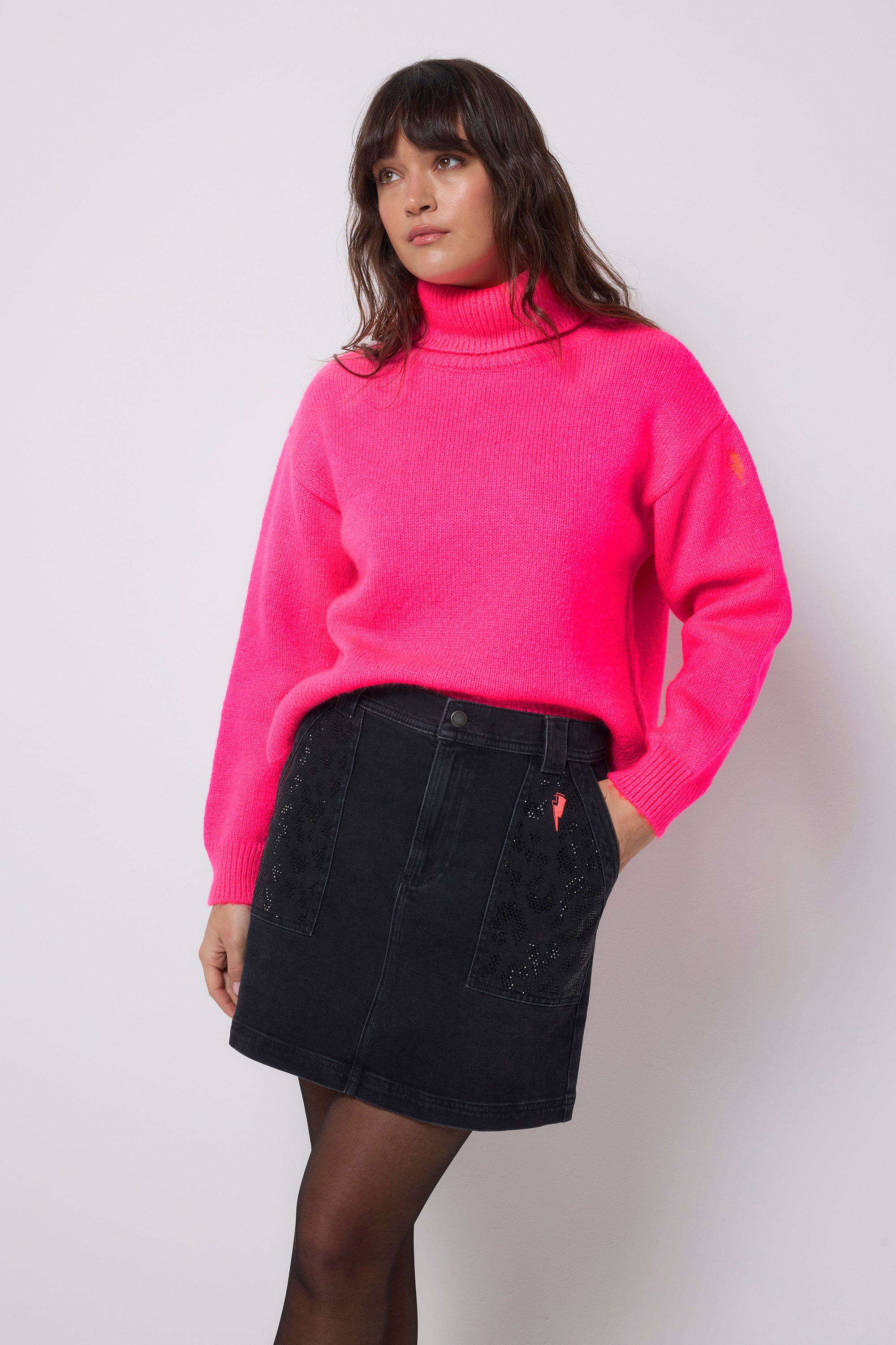 Scamp and Dude Washed Black Embellished Leopard Patch Pocket Denim Mini Skirt | Model wearing a bright pink knit jumper with a black denim mini skirt that features embellished leopard print detail on the pockets.
