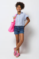 Scamp & Dude Mid Indigo Denim Shorts | Model holding a pink bag and wearing a striped shirt and blue denim shorts and pink trainers on a white background.