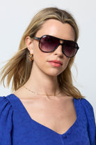 Scamp and Dude Black Aviator Sunglasses | Model wearing a blue broderie dress with gold jewellery and a pair of aviator style sunglasses in a black colour.