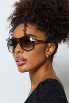 Scamp and Dude Tortoiseshell Aviator Sunglasses | Model wearing a black dress with gold jewellery and a pair of aviator style sunglasses in a tortoiseshell colour.