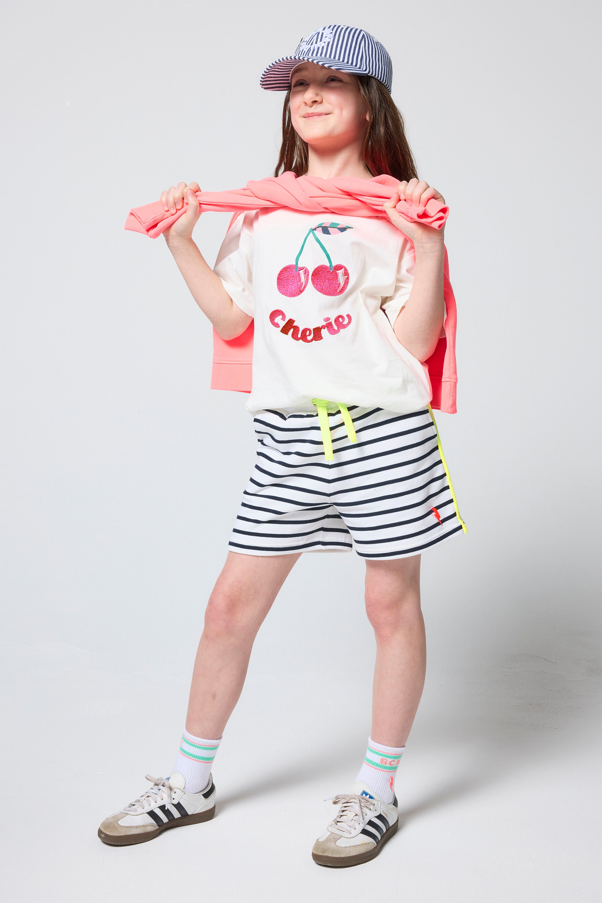 Scamp & Dude Kids White Cherry Print T-Shirt | Model wearing a cherry-print shirt, striped shorts, and a cap.