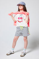 Scamp & Dude Kids White Cherry Print T-Shirt | Model wearing a cherry-print shirt, striped shorts, and a cap.