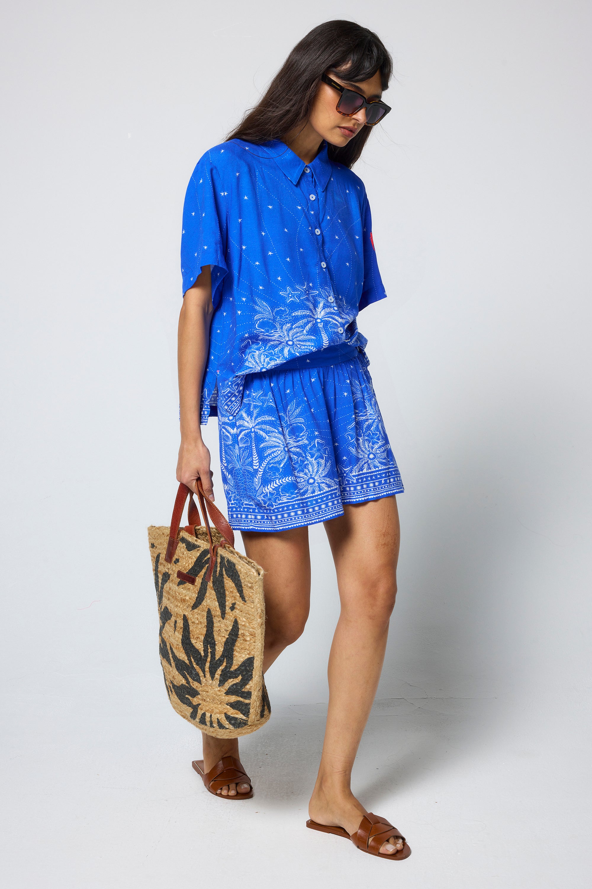Scamp & Dude Blue & White Sundream Tropical Placement Print Shorts | Model wearing a blue shirt and shorts with a white tropical pattern, holding a woven bag.