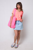 Scamp & Dude Washed Neon Coral V-Neck T-Shirt | Model wearing a bright coral v-neck t-shirt with a blue denim skirt and blue trainers. Holding a neon pink quilted tote bag.