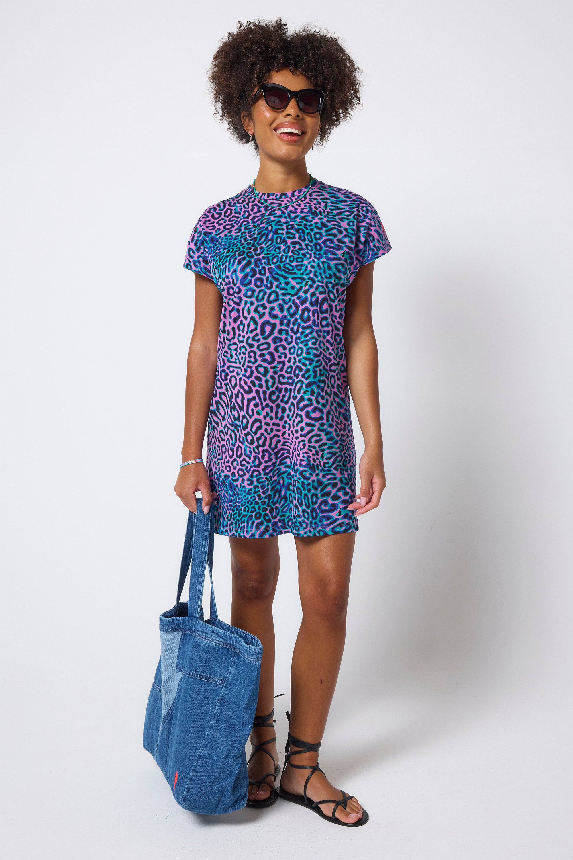 Scamp & Dude Pink with Green Leopard Ray T-Shirt Dress | Model wears a short t-shirt dress in a pink and green leopard ray print. Worn with black strappy sandals and a denim tote bag.