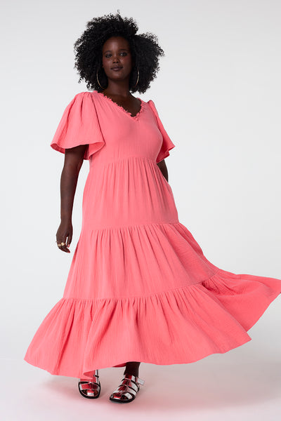 Scamp and Dude Coral Flute Sleeve Tiered Midi Dress | Model wearing a coral v neck midi dress featuring flute sleeves and tiered skirt detail. Paired with metallic look sandals.