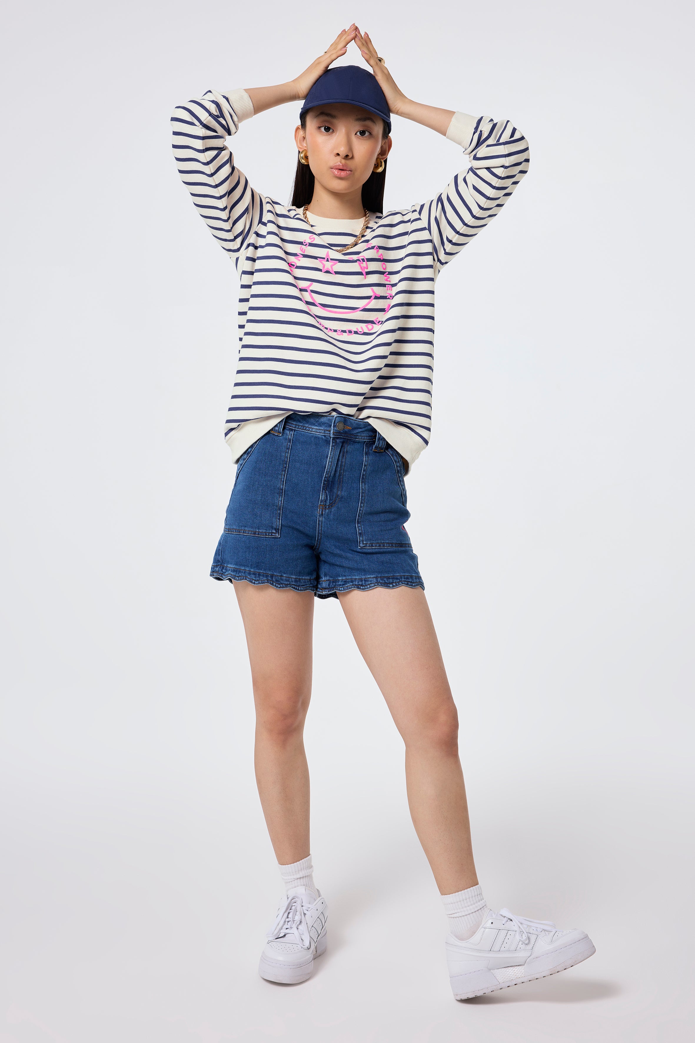 Scamp and Dude Ivory with Blue Stripe and Smiley Face Oversized Sweatshirt | Model wearing a round neck sweatshirt featuring stripes and smiley face, worn with scallop hem denim shorts and a navy cap.