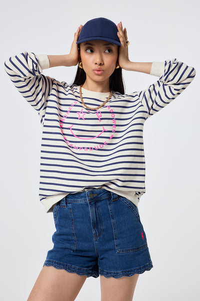 Scamp and Dude Ivory with Blue Stripe and Smiley Face Oversized Sweatshirt | Model wearing a round neck sweatshirt featuring stripes and smiley face, worn with scallop hem denim shorts and a navy cap.