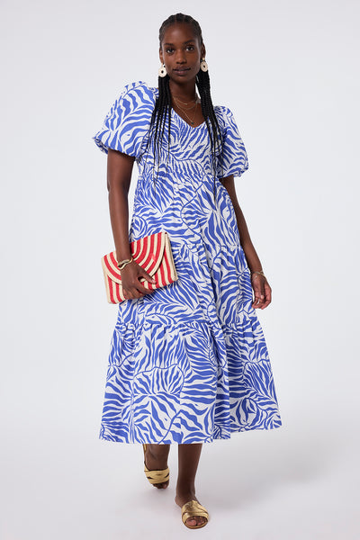 Scamp and Dude White with Blue Palm Shirred Puff Sleeve Maxi Dress | Model wearing white puff sleeve maxi dress with blue palm and lightning bolt print paired with gold sandals and a straw clutch bag.