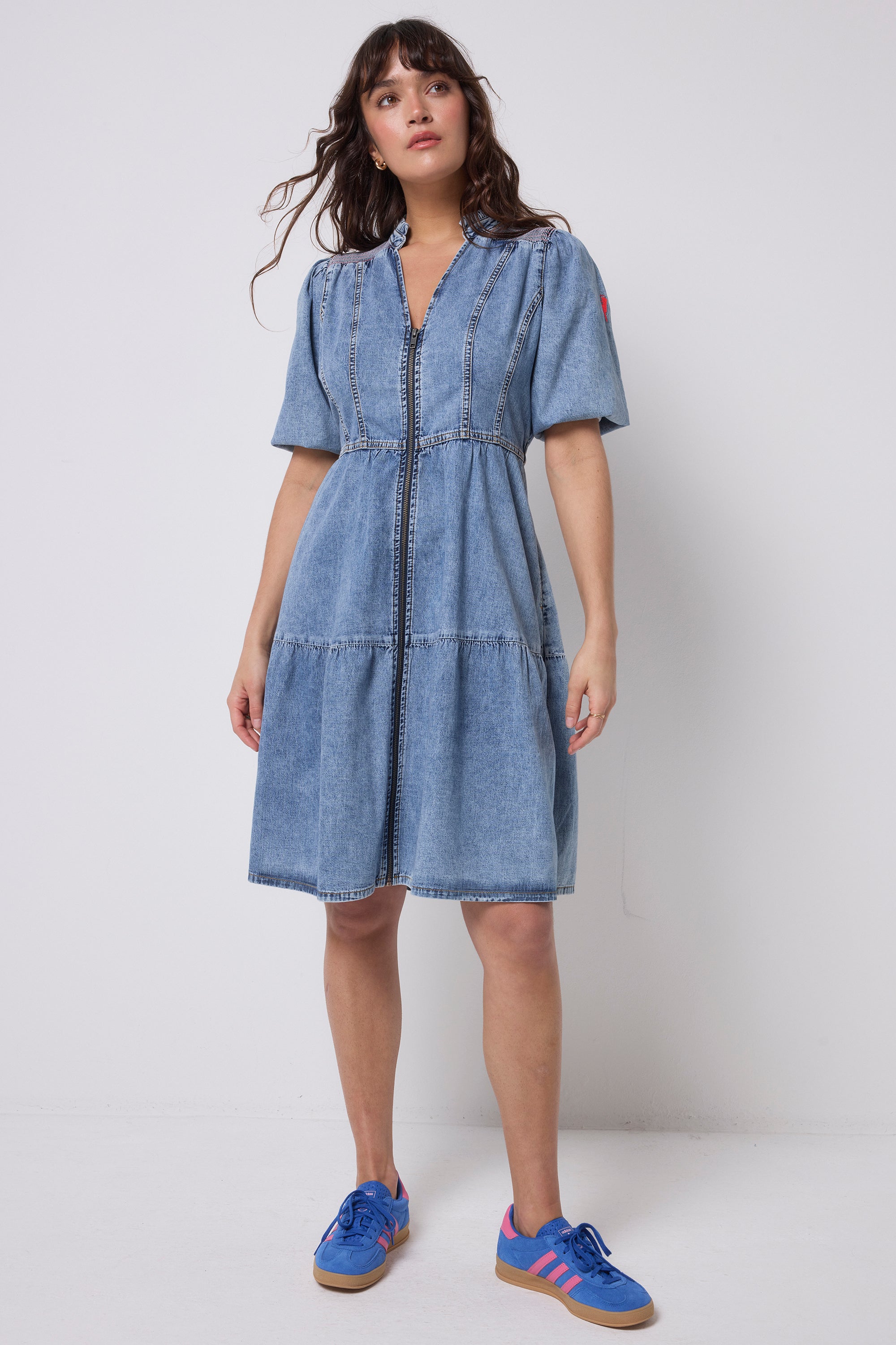 Scamp and Dude Pale Indigo Zip Detail Tiered Short Denim Dress | Model wearing a short pale blue denim dress with zip front and tiered detail. Worn with blue trainers.