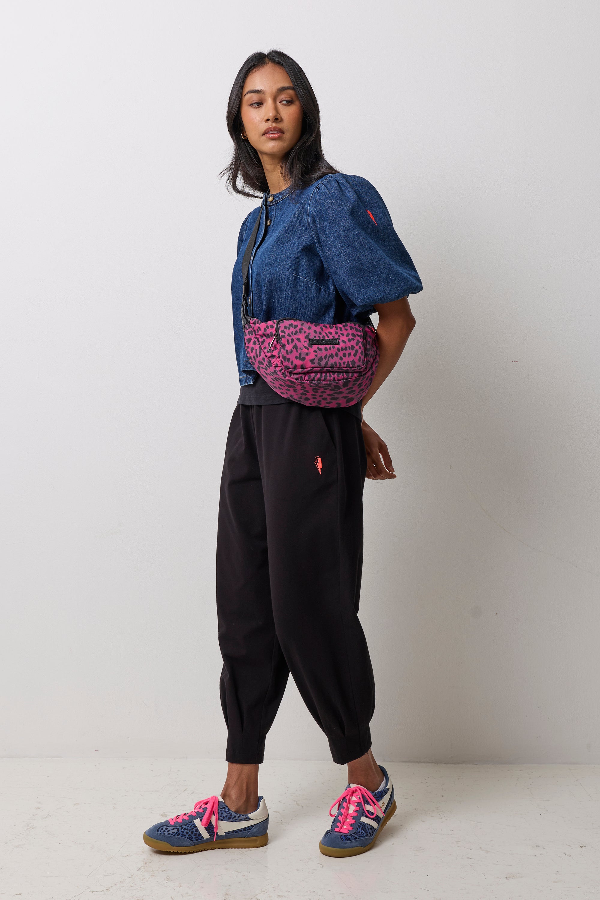 Scamp & Dude Black Cherry Cheetah Bum Bag | Model wearing a blue cropped jacket with puff sleeves, black jogger pants, and blue sneakers with bright pink laces. She is carrying a pink and black leopard-print bum bag.