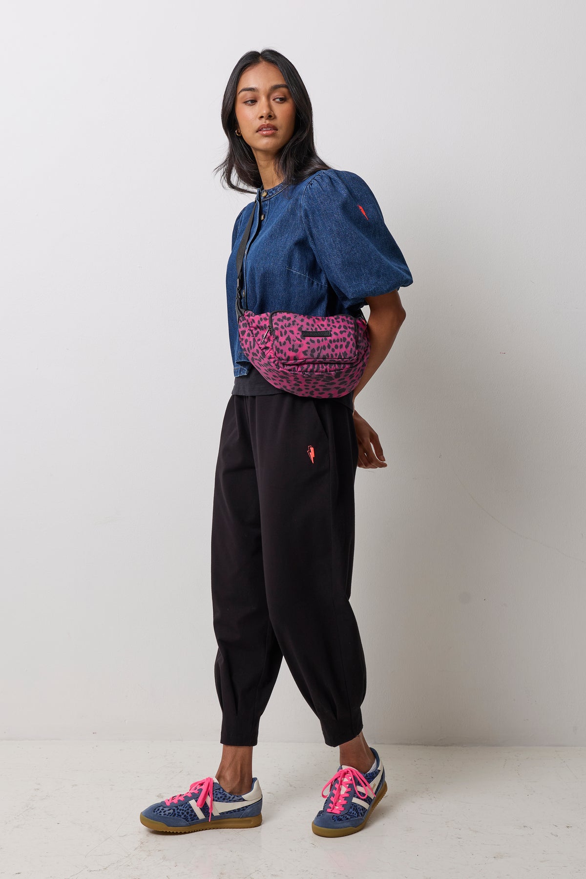 Scamp & Dude Black Cherry Cheetah Bum Bag | Model wearing a blue cropped jacket with puff sleeves, black jogger pants, and blue sneakers with bright pink laces. She is carrying a pink and black leopard-print bum bag.