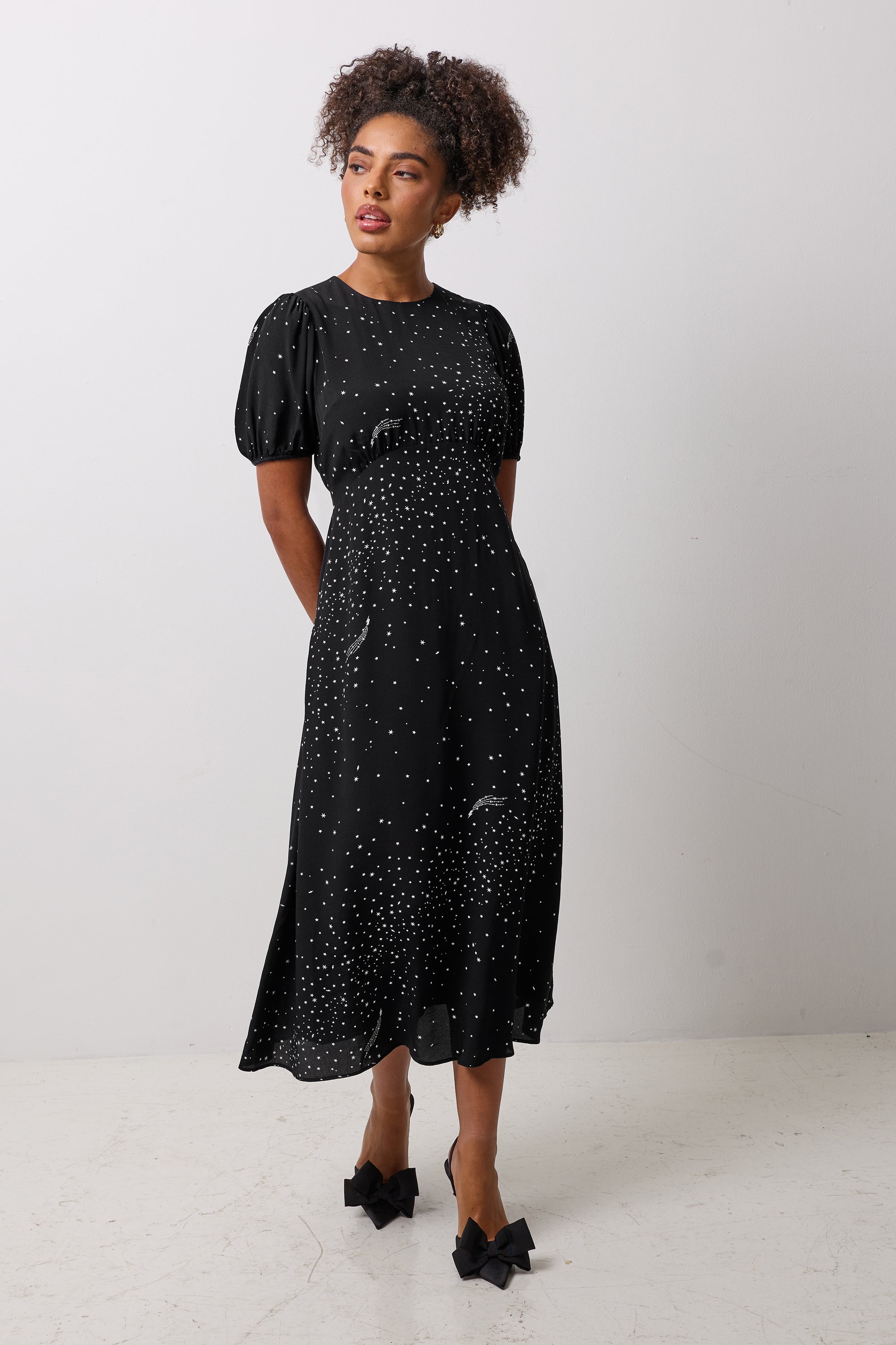 Scamp & Dude Black with White Written In The Stars Print Curved Waist Midi Dress | Model wearing a black midi dress with white star print and black bow detail high heels.