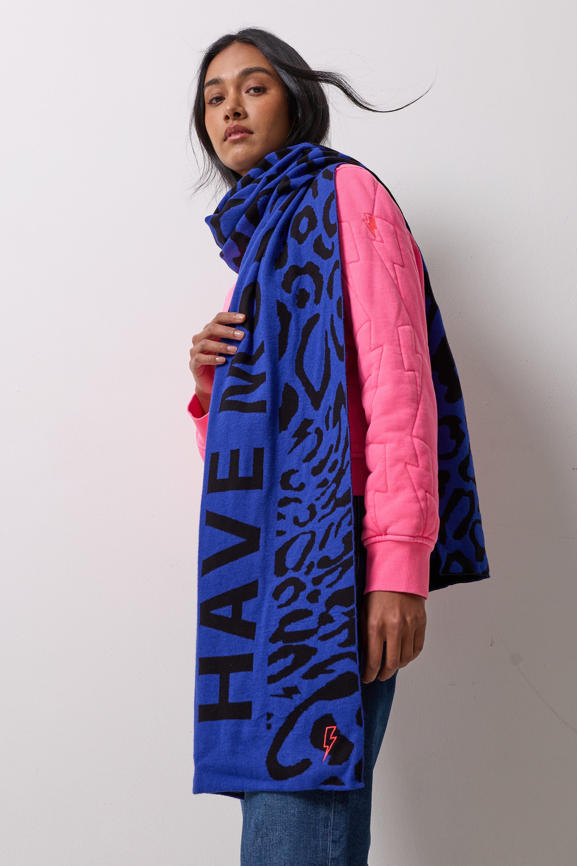 Scamp and Dude Navy with Black Slogan Jacquard Scarf | Model wearing a pair of blue jeans and a pink bomber jacket with a blue and black printed scarf. 