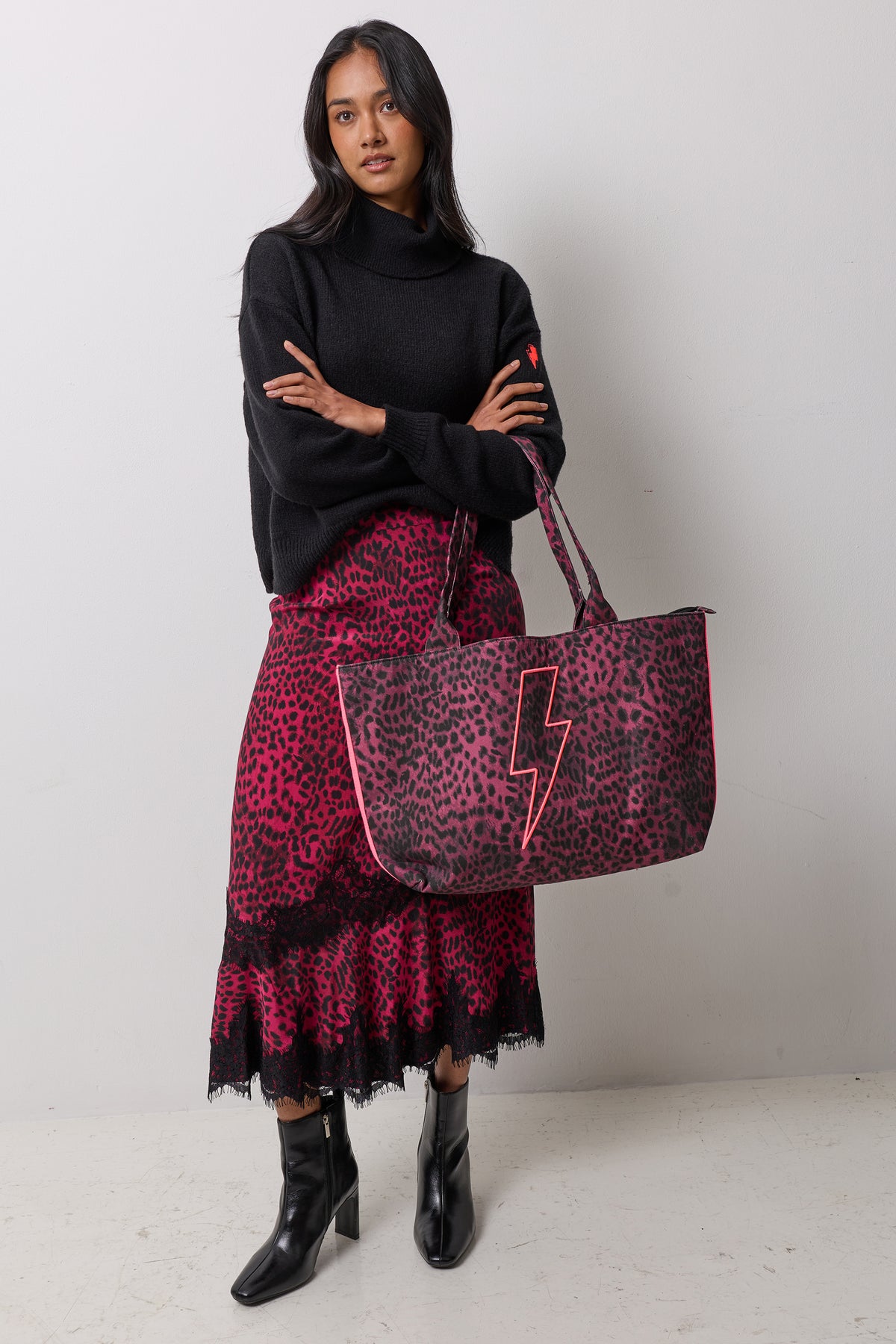 Scamp & Dude Black Cherry Cheetah Canvas Shopper Bag | Model wearing a black turtleneck sweater, a pink and black leopard-print skirt, and black ankle boots, while holding a large matching pink and black leopard-print tote bag with a pink lightning-bolt detail.