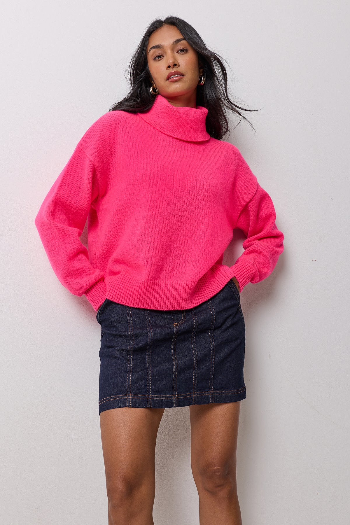 Scamp & Dude Neon Pink Roll Neck Knitted Jumper | Model wearing a bright pink turtleneck knit sweater paired with a dark denim mini skirt.